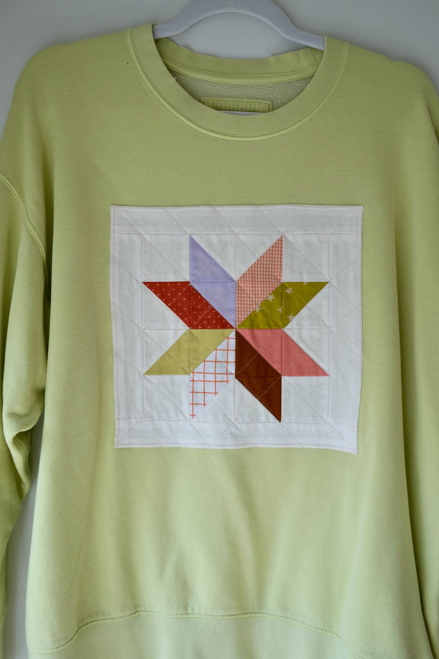 A pale yellow-green crewneck sweatshirt featuring a square white patchwork chest design with a multi-colored star quilted pattern.