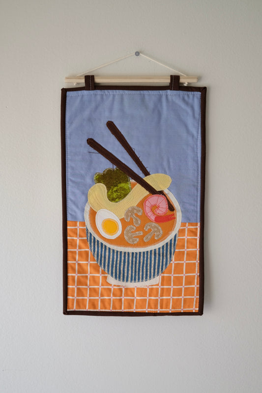 A rectangular fabric wall hanging with a stylized ramen bowl illustration, suspended from a wooden dowel. 