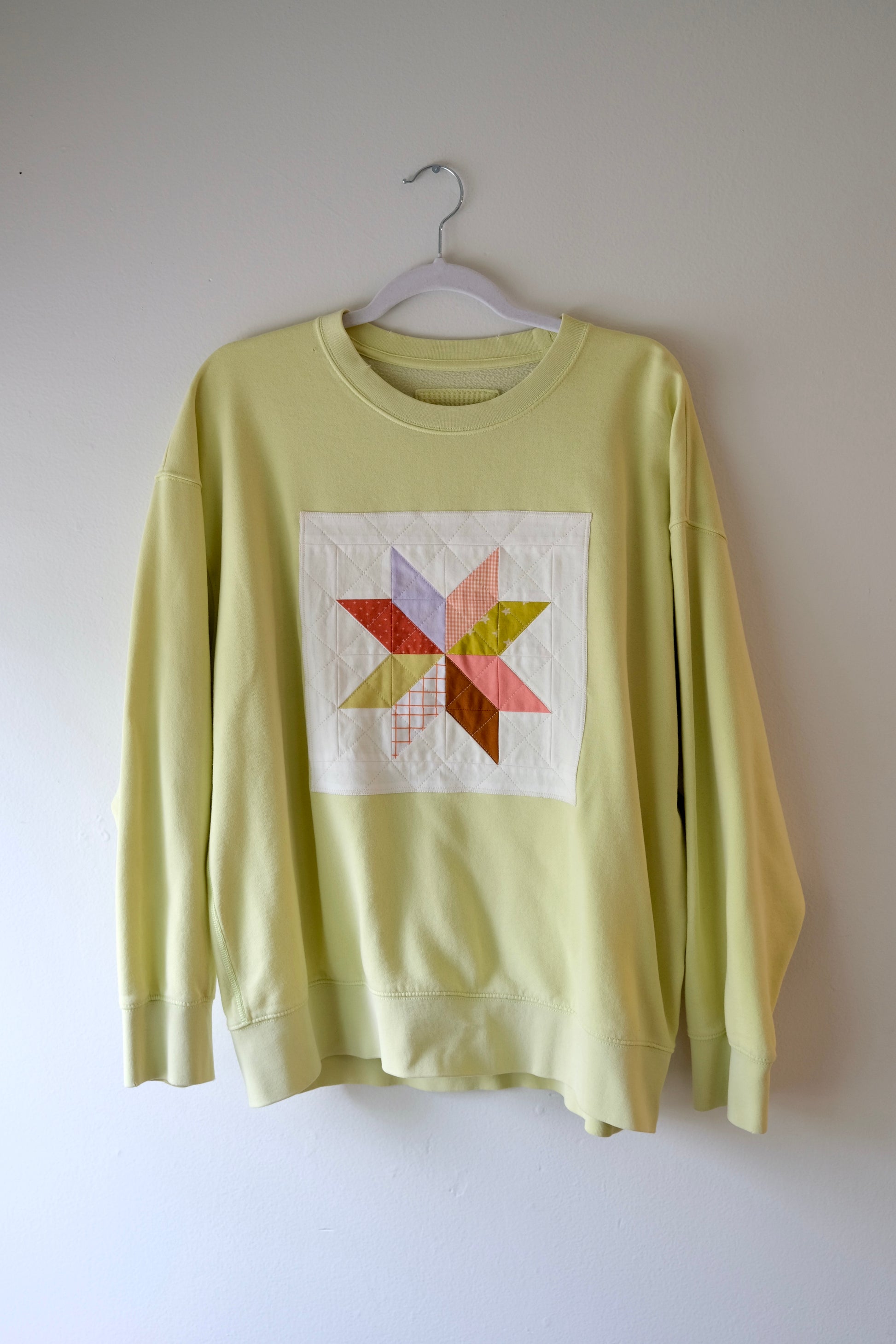 A pale yellow-green crewneck sweatshirt. The sweatshirt features a square white patchwork graphic on the chest with a multi-colored star design.